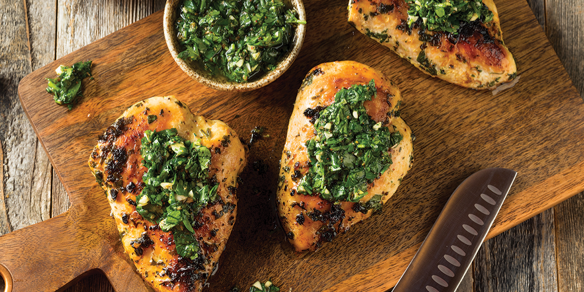 Grilled chicken breasts with green herb sauce on a wooden cutting board.
